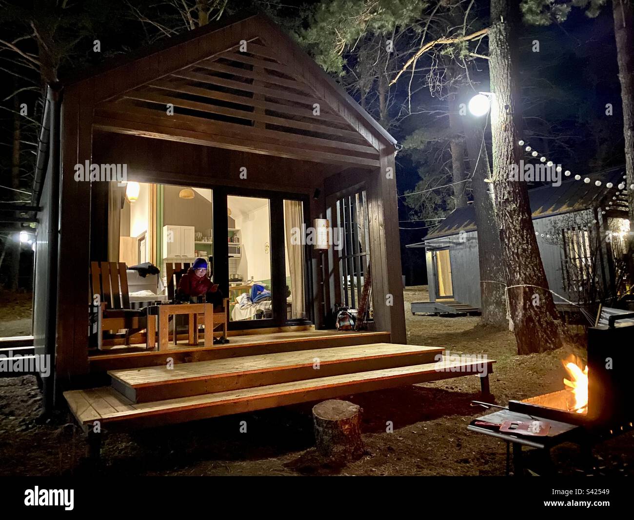 A girl is sitting on the veranda of a barnhouse-style cabin at night behind the light of a glass door and lanterns in the woods. - Smartphone Captured Stock Image