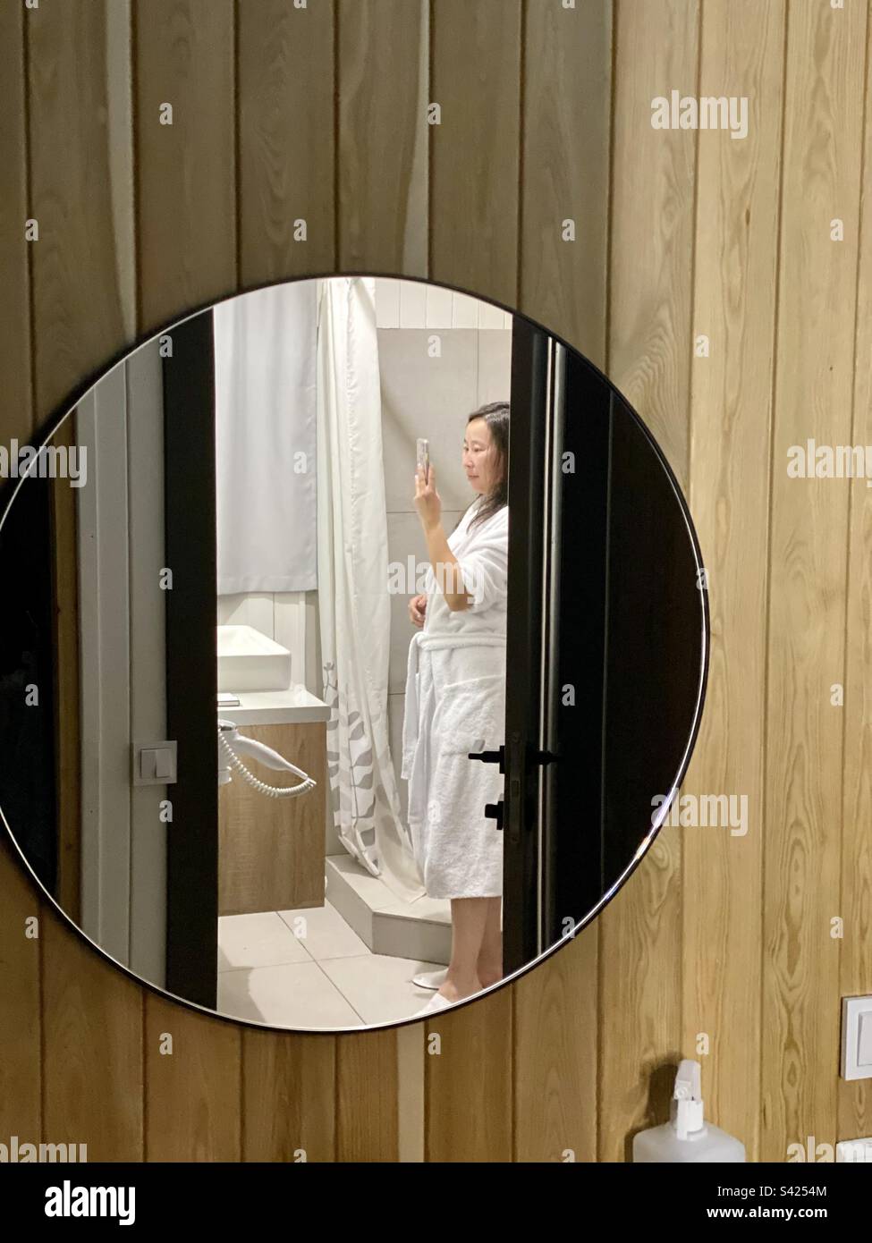 Reflection in a round mirror of a girl in a white coat in a bathtub taking a selfie. Stock Photo