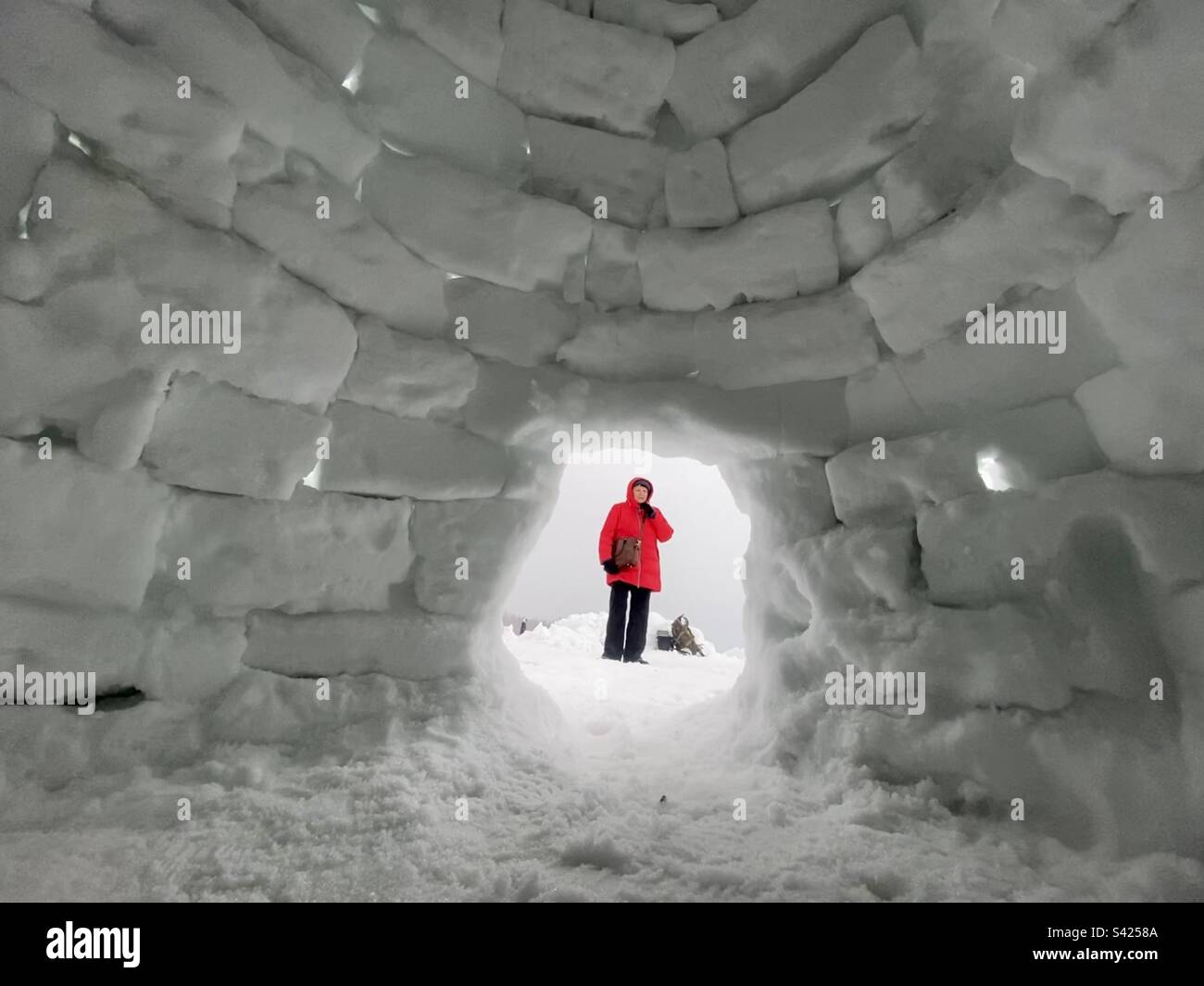 A woman in a bright jacket stands against the background of a brick igloo exit from a snowy house. - Smartphone Captured Stock Image