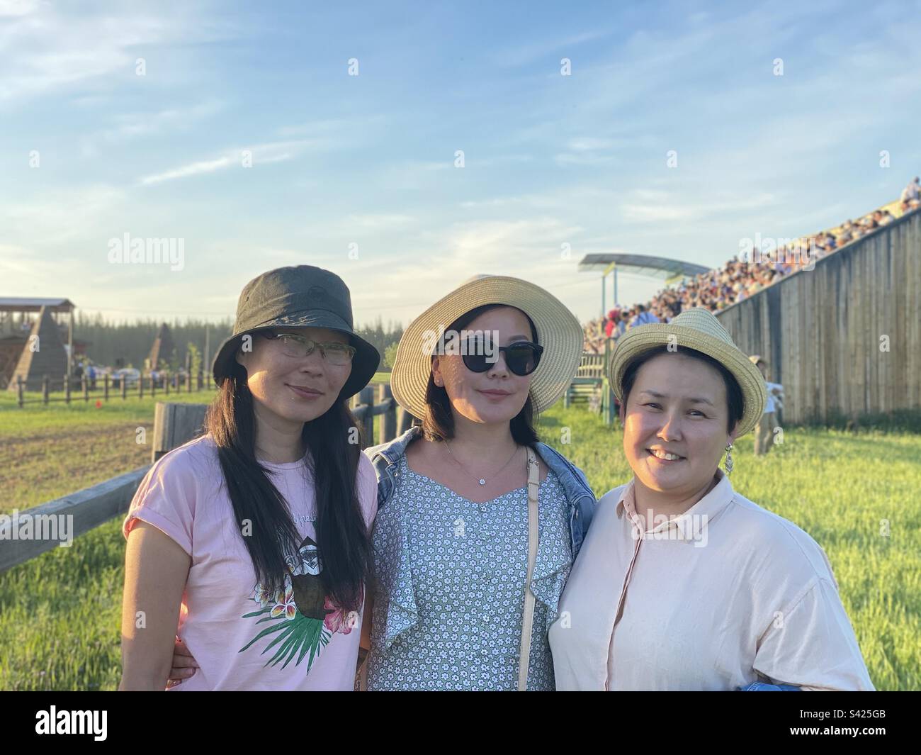 Three friends of Asian Yakut girls smile against the background of the stands in the summer at a holiday in Yakutia. Stock Photo