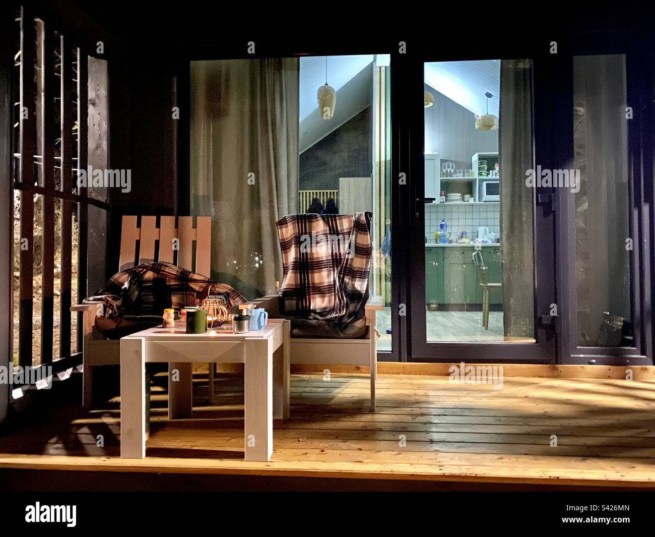 The exterior of the cottage with a veranda and barnhouse-style glass doors, at night with lights and chairs with a blanket. - Smartphone Captured Stock Image
