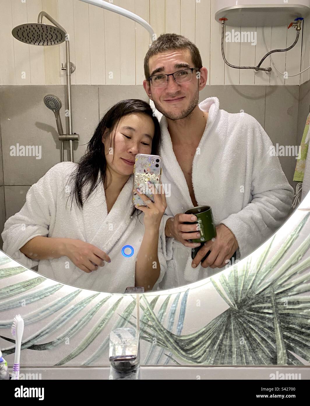 An Asian girl and a European man in white coats take a selfie on an iPhone 11 in a bathtub in a mirror. - Smartphone Captured Stock Image