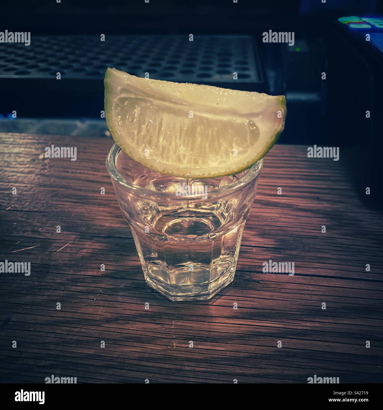 A photograph of a tequila shot with a slice of line on top of the glass, on a wooden bar. - Smartphone Captured Stock Image
