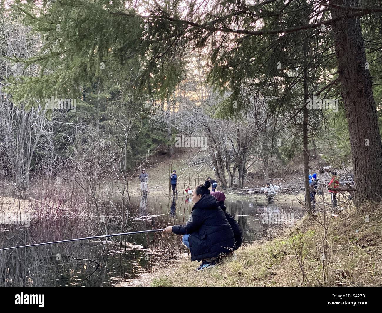 A lot of young fishermen with fishing rods of Yakut women, men and children are fishing on the little taiga river in the spruce forest Stock Photo
