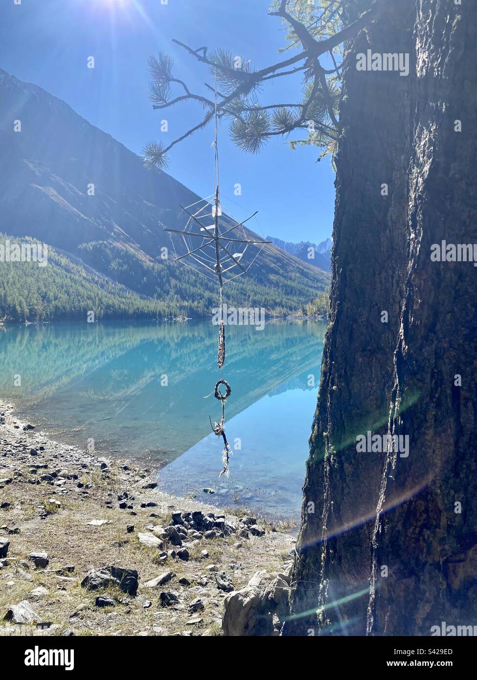 A dreamcatcher hangs on a tree in the sunlight in the forest in front of the mountains behind the lake in Altai in Siberia. - Smartphone Captured Stock Image