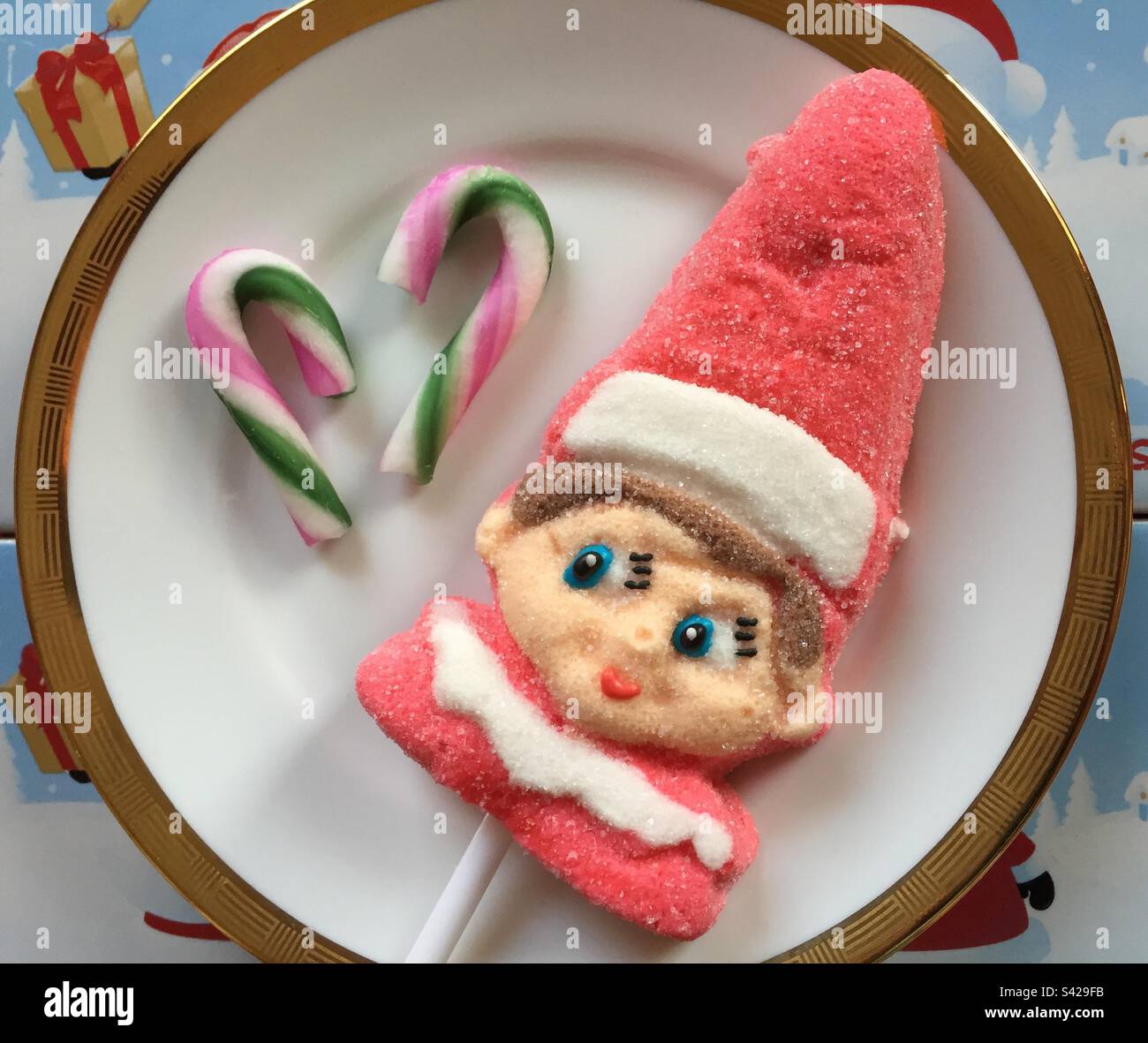 Christmas Elf lollipop, appearing to look at a couple of candy canes on the same plate. Brightly coloured festive sweets made of sugar. Pic 4. - Smartphone Captured Stock Image