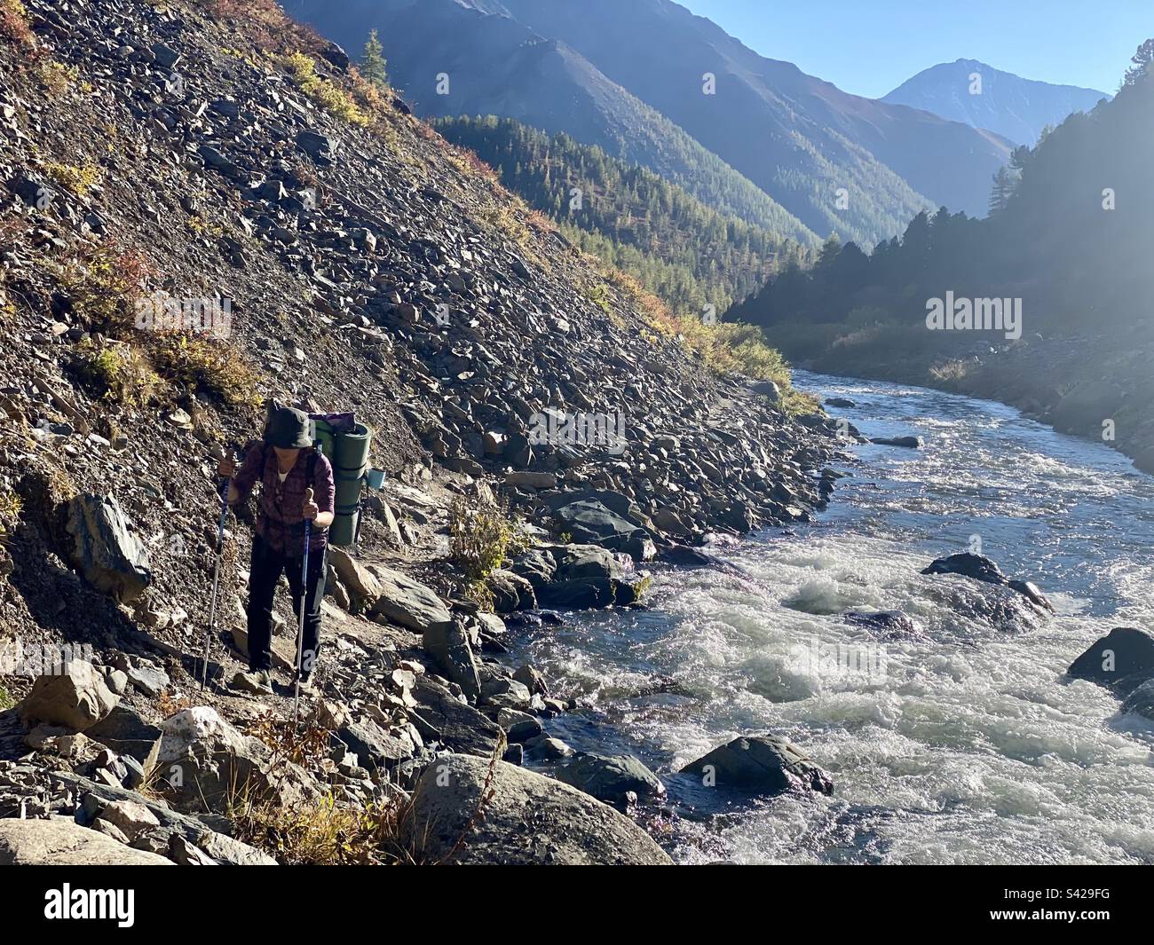 A traveler girl with a backpack walks along a narrow path under a rock along a mountain river in the Altai in Siberia. - Smartphone Captured Stock Image