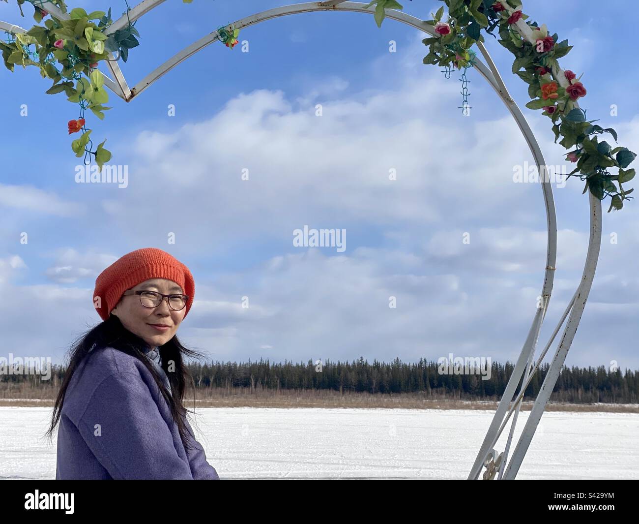 A Yakut Asian girl is sitting half a turn in spring clothes on a bench in the form of a heart with flowers by the ice of the snow of the river. Stock Photo