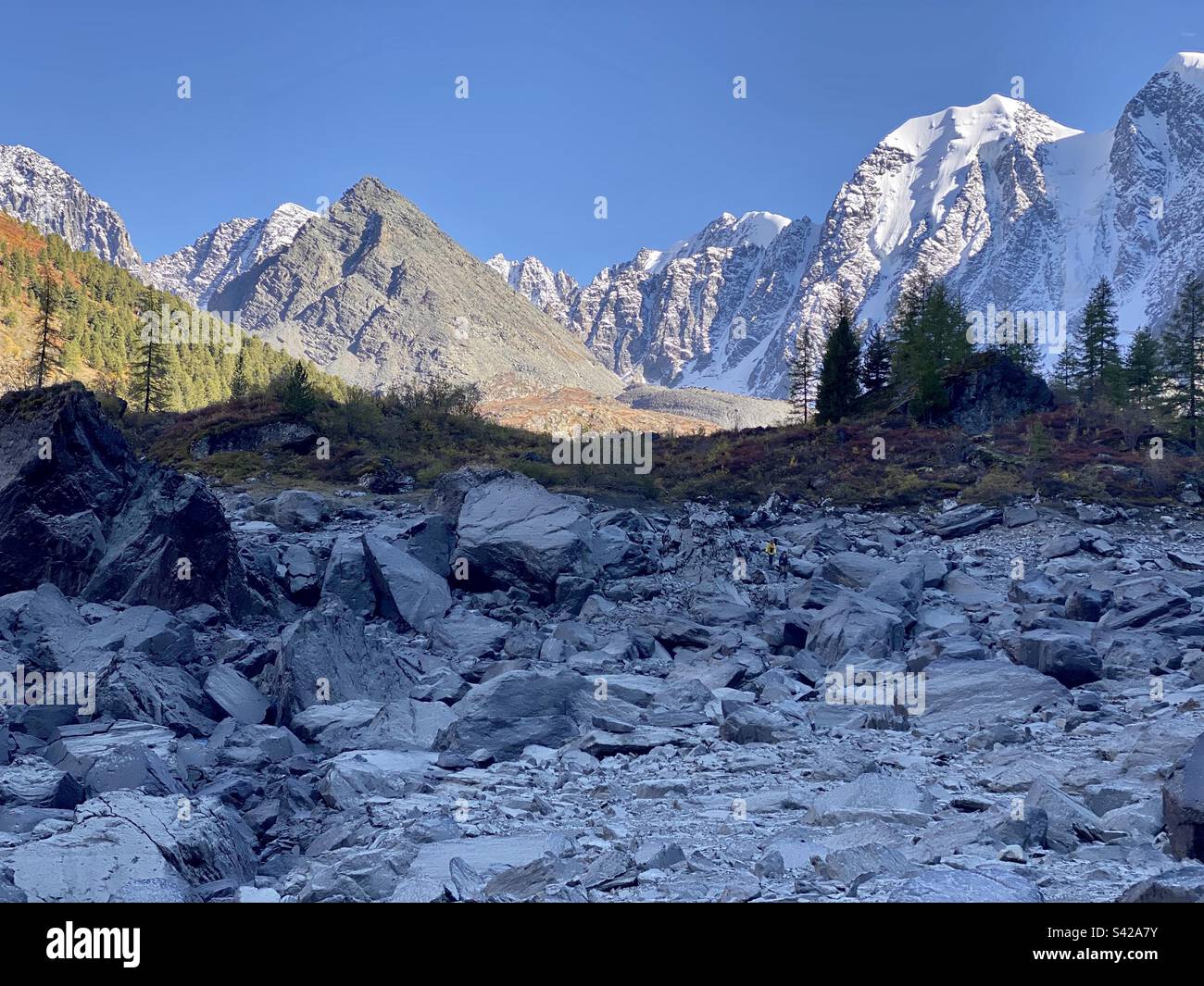 A couple of tourists in the distance are walking on the stones of a dried-up lake under mountains with glaciers and snow in the Altai in Siberia. - Smartphone Captured Stock Image