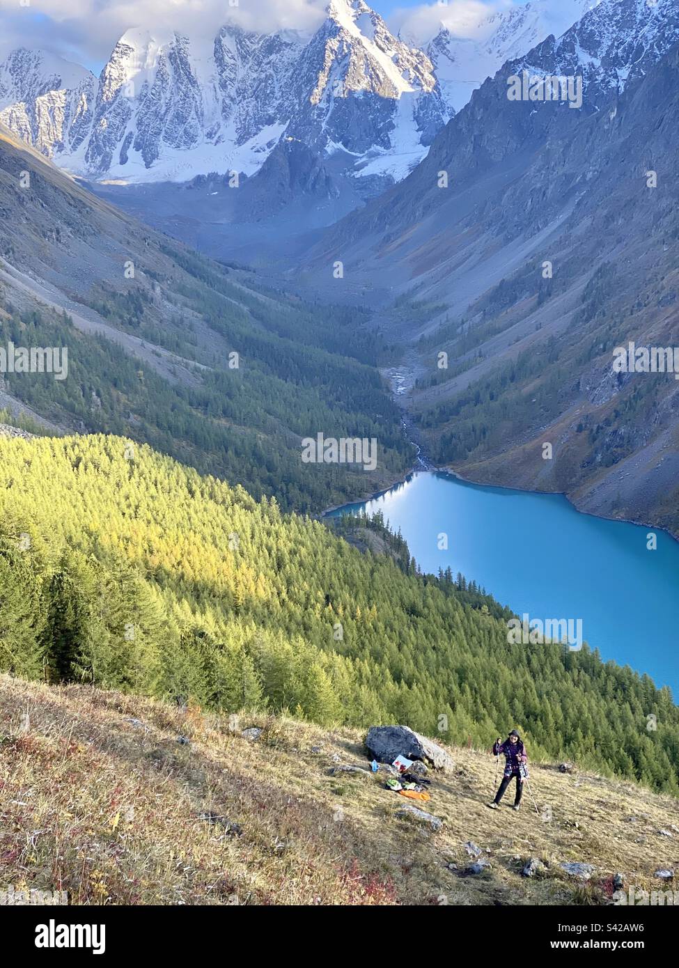 A Yakut Asian girl traveler stands smiling on a hill above an alpine lake and mountains with glaciers and snow in the Altai. - Smartphone Captured Stock Image