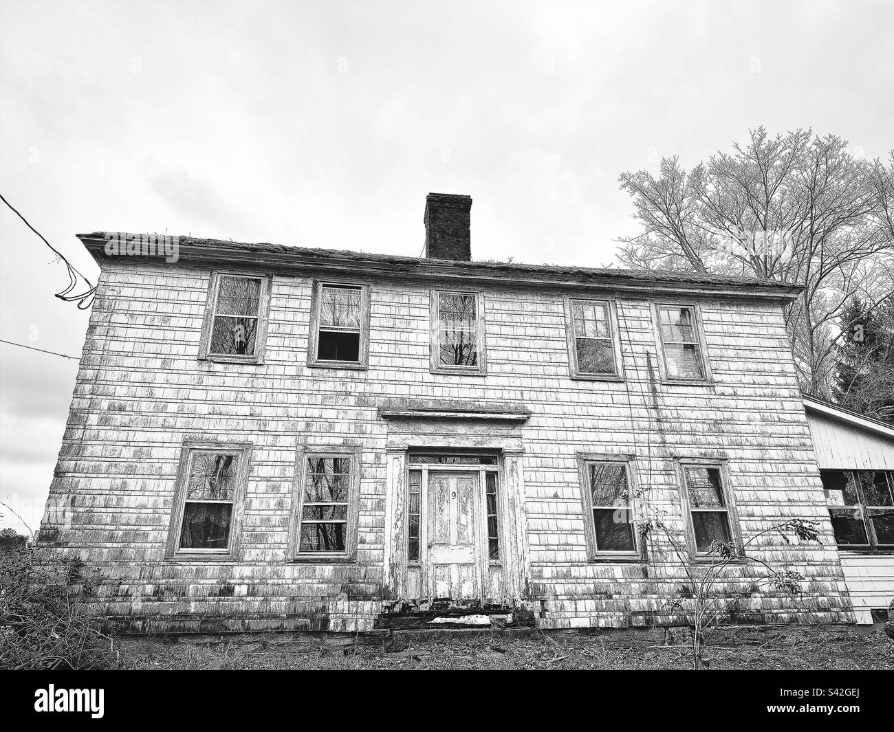 Black-and-white photo of an old, rundown white farmhouse in Lebanon, Connecticut during winter. The exterior looks dirty, and is deteriorating. Stock Photo