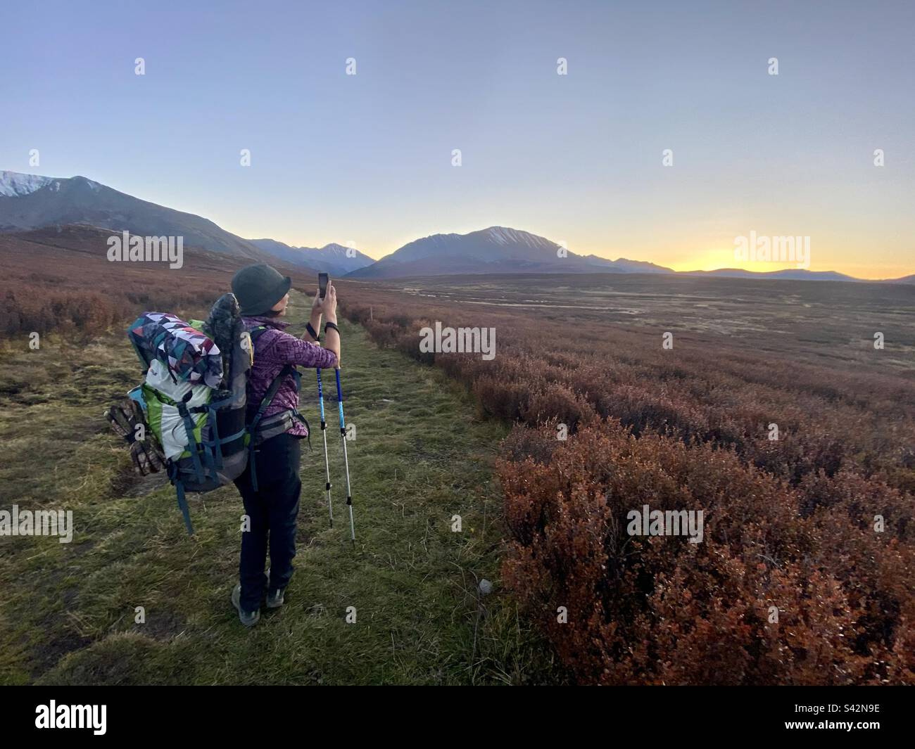 A traveler girl with a large backpack takes photos on her phone of a sunset on a pass among the mountains in Altai in Russia. - Smartphone Captured Stock Image