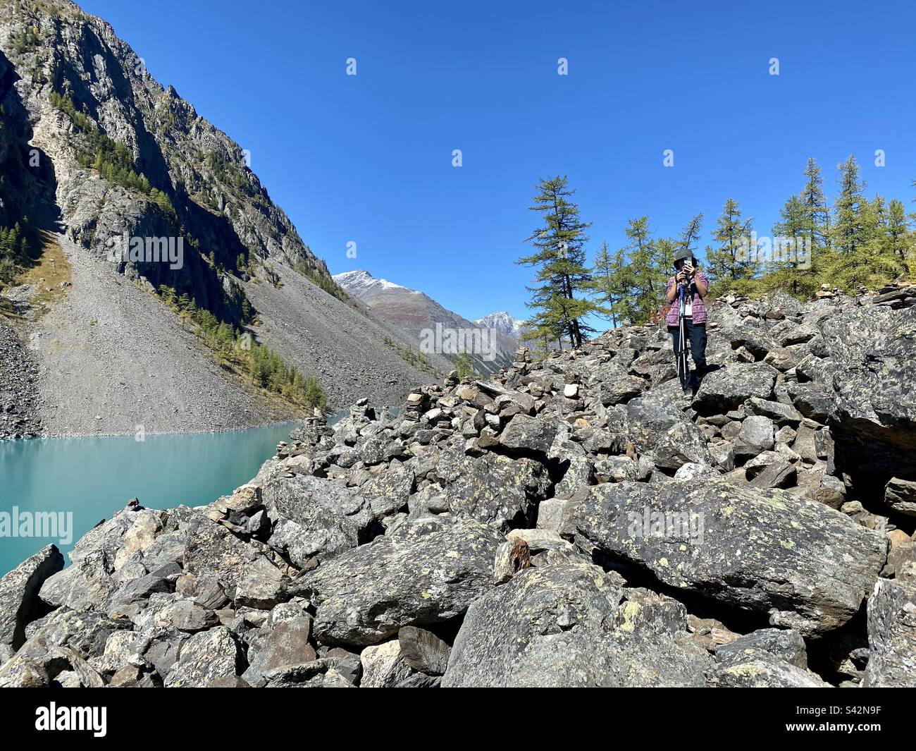 A girl traveler takes pictures on her phone of large stones at an alpine lake among the mountains in the Altai in Russia. Stock Photo