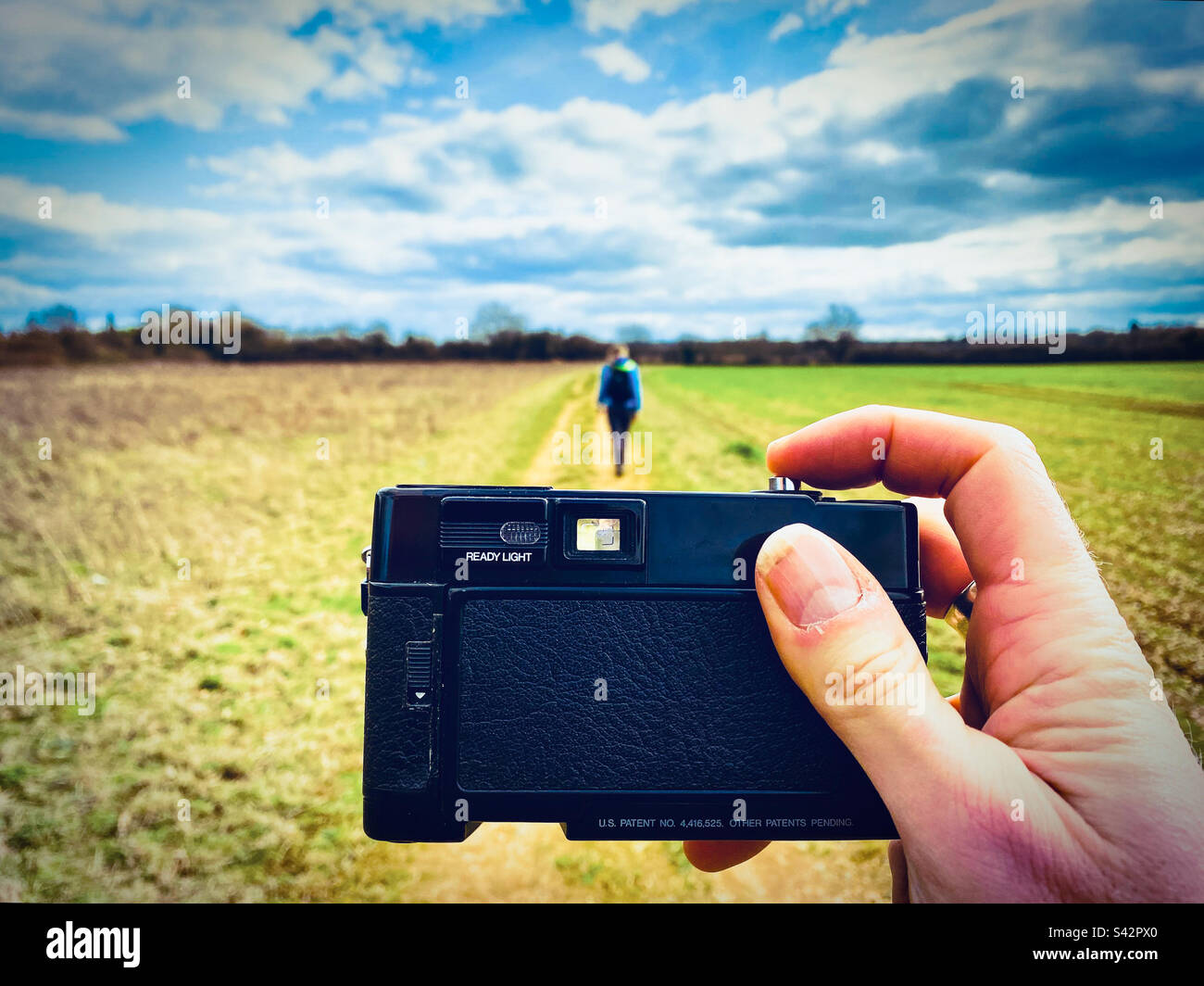A hand holds a retro film camera taking a picture of a person walking in the countryside in England UK Stock Photo