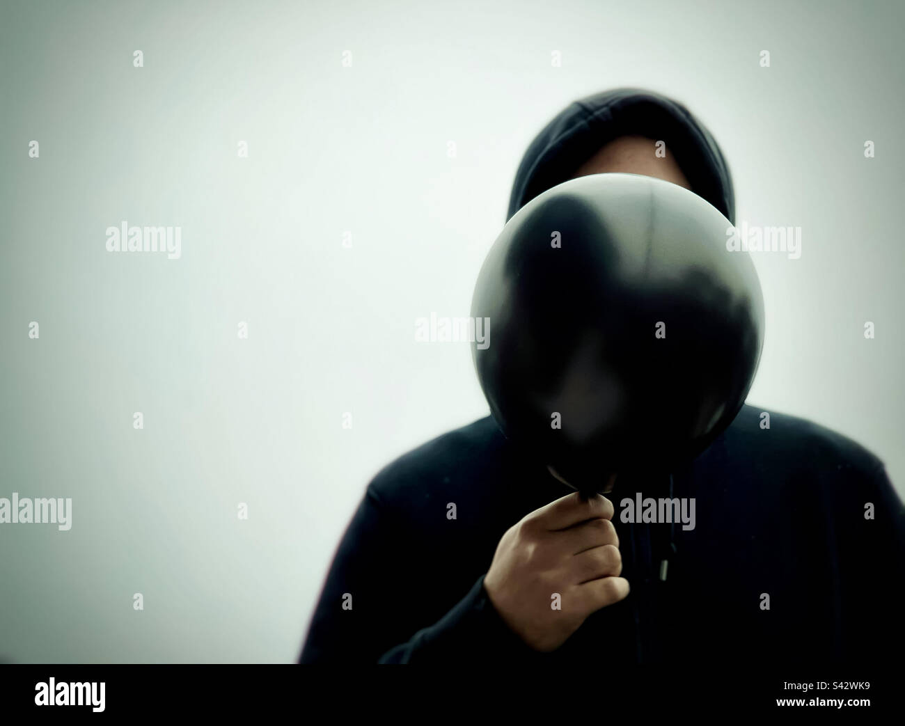 Portrait of man in black hoodie holding a black balloon in front of his face against white background. - Smartphone Captured Stock Image