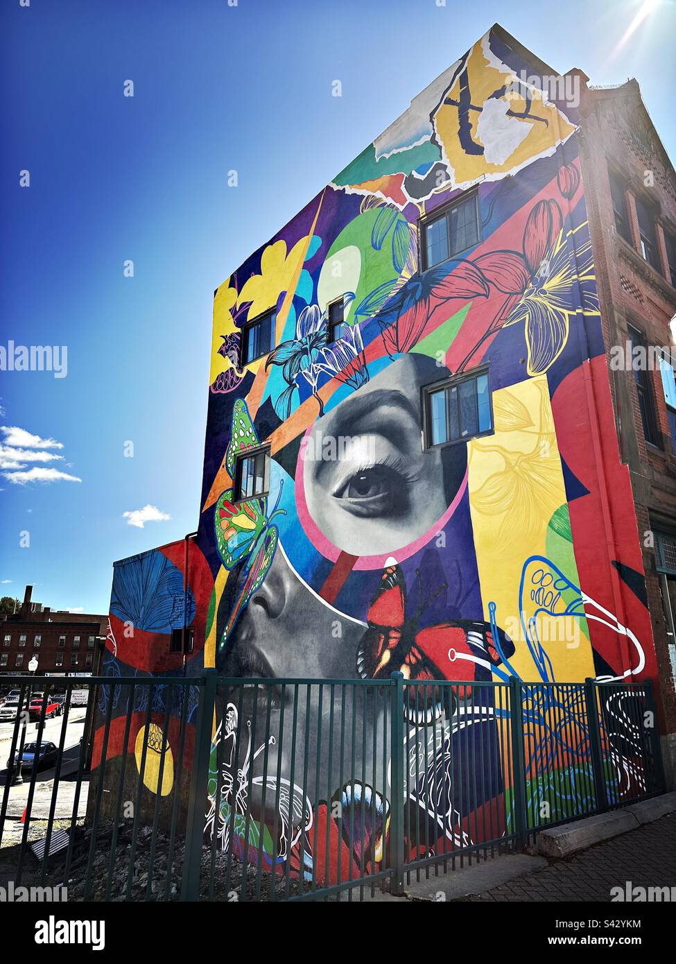 Streetscape with wall mural art and black metal fence in Norwich, Connecticut, USA. With blue sky and a ray of sun shining down from the top right corner. - Smartphone Captured Stock Image