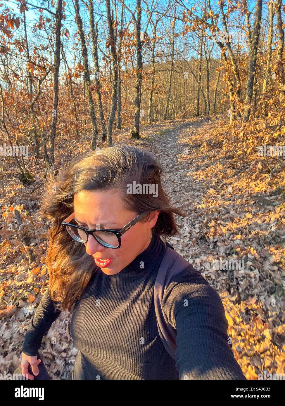 Woman with eyeglasses, long brown hair, brown skin and a black turtleneck shirt walking, hiking on a trail with dry leaves, in the autumn, during golden hour. Sun reflecting at the leaves and blue sky Stock Photo