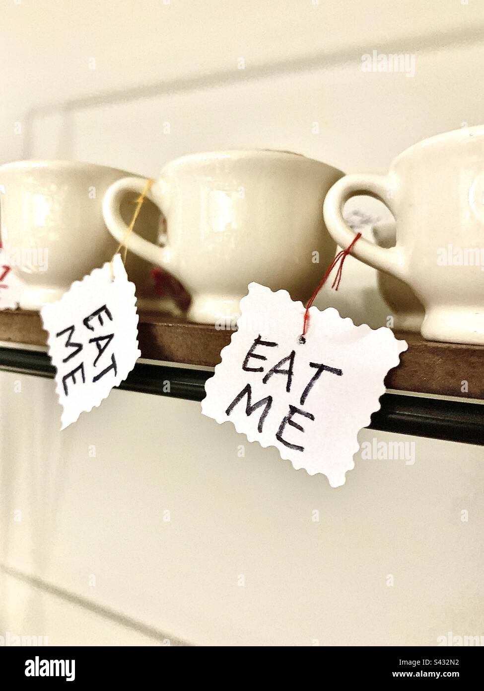 Three Alice in Wonderland themed cupcakes inside of white teacups on wooden shelf against white background. Cups are labeled with the words, EAT ME. Includes copy space. - Smartphone Captured Stock Image