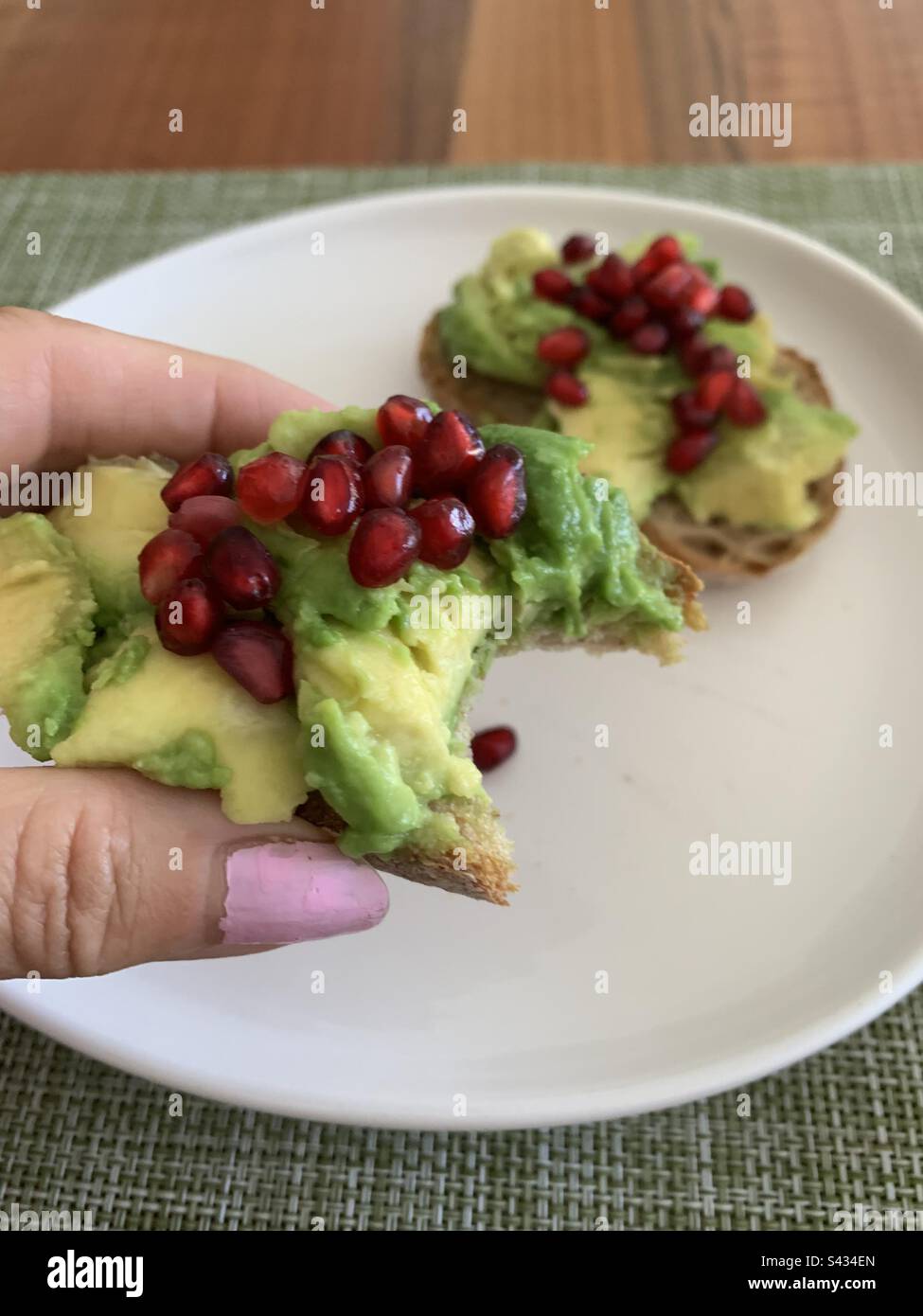 Closeup image of a woman holding a half eaten slice of sourdough avocado toast topped with pomegranate arils - Smartphone Captured Stock Image