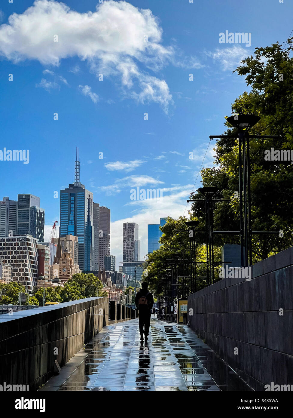 Rear view of man walking along paved footpath against buildings and city skyline after the rain in Melbourne, Australia. - Smartphone Captured Stock Image Rear view of man walking along paved footpath against buildings and city skyline after the rain in Melbourne, Australia. - Smartphone Captured Stock Image