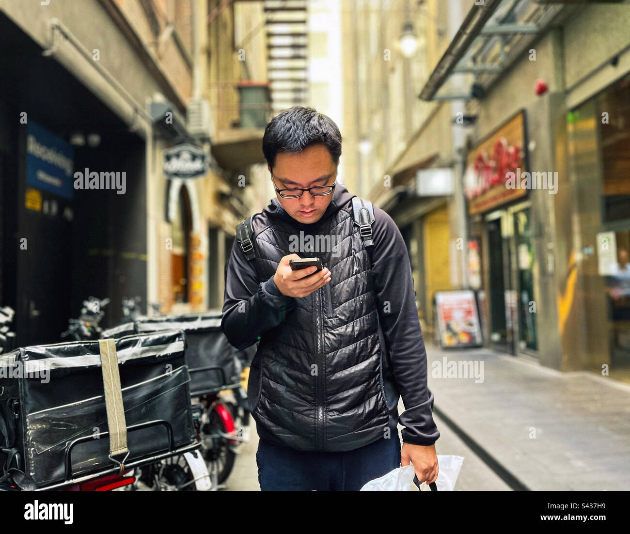 Young Asian man using smartphone while standing beside food delivery bicycles in laneway against buildings in the city in Melbourne, Australia. - Smartphone Captured Stock Image