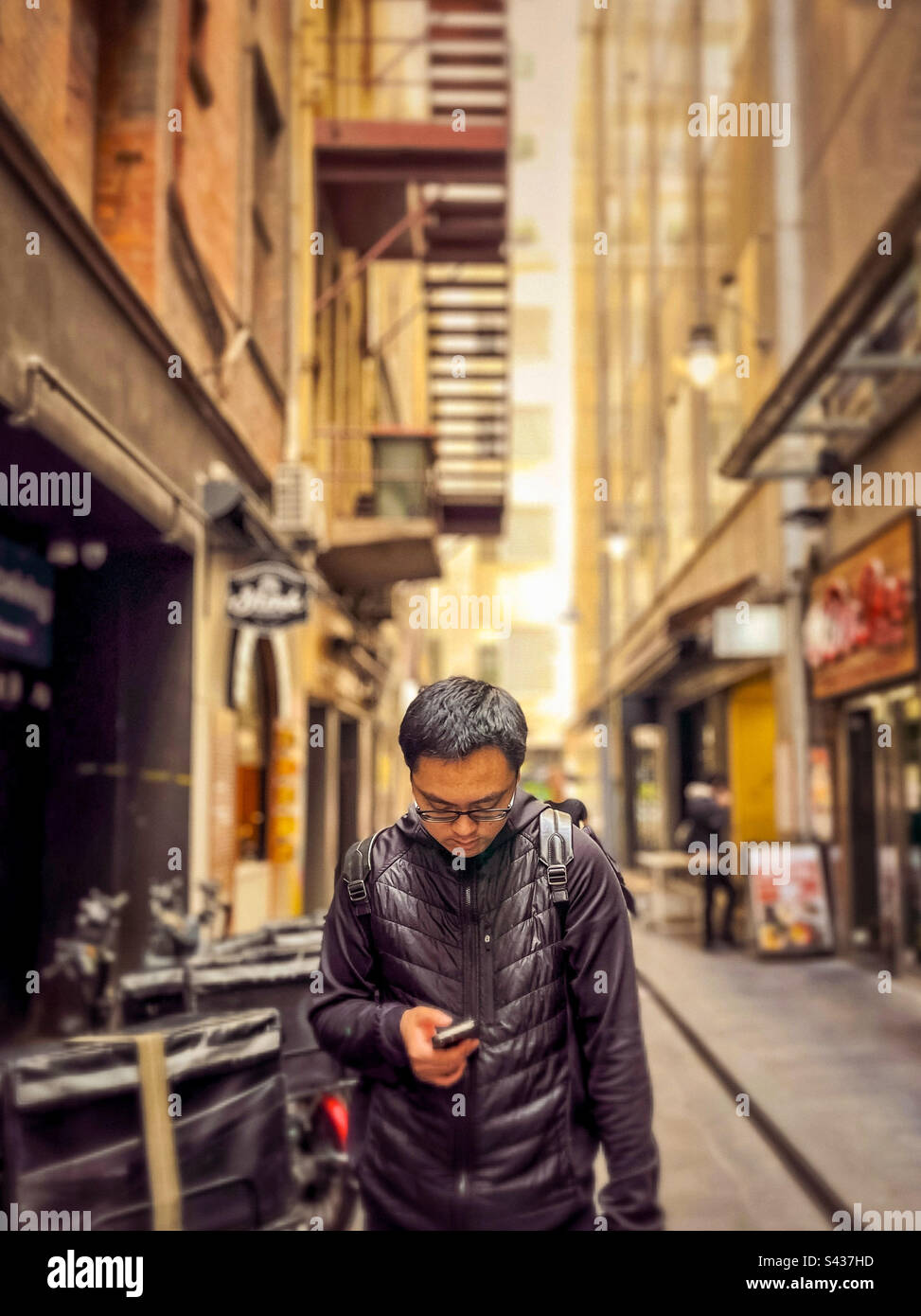 Young Asian man using mobile phone while standing beside food delivery bicycles in laneway against buildings in the city in Melbourne, Australia. - Smartphone Captured Stock Image