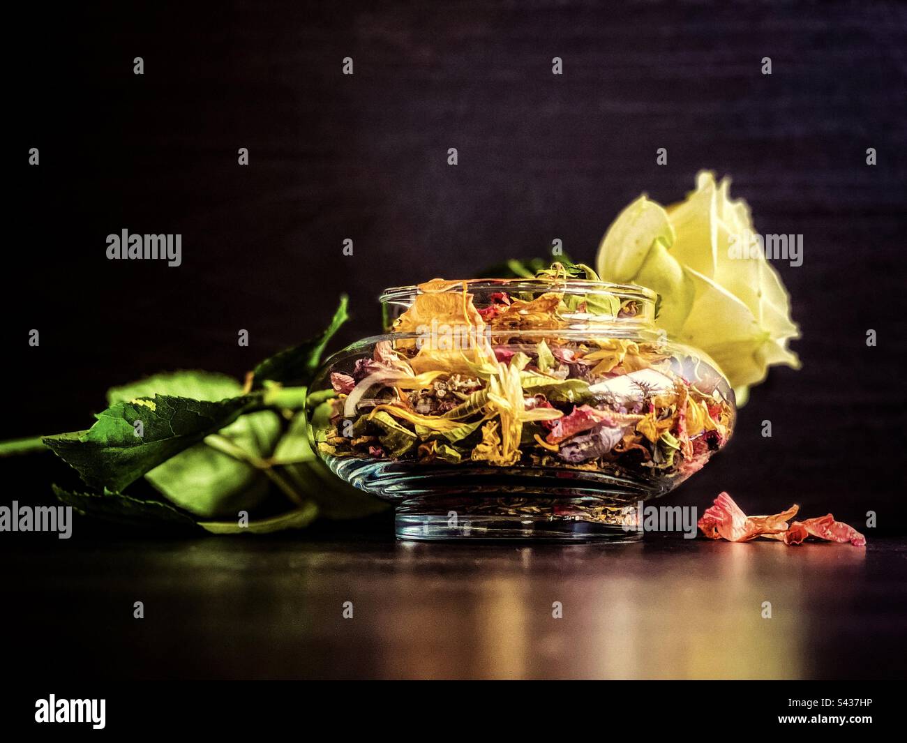 Close-up of single yellow rose and potpourri in glass container with dried flower petals on black background. - Smartphone Captured Stock Image