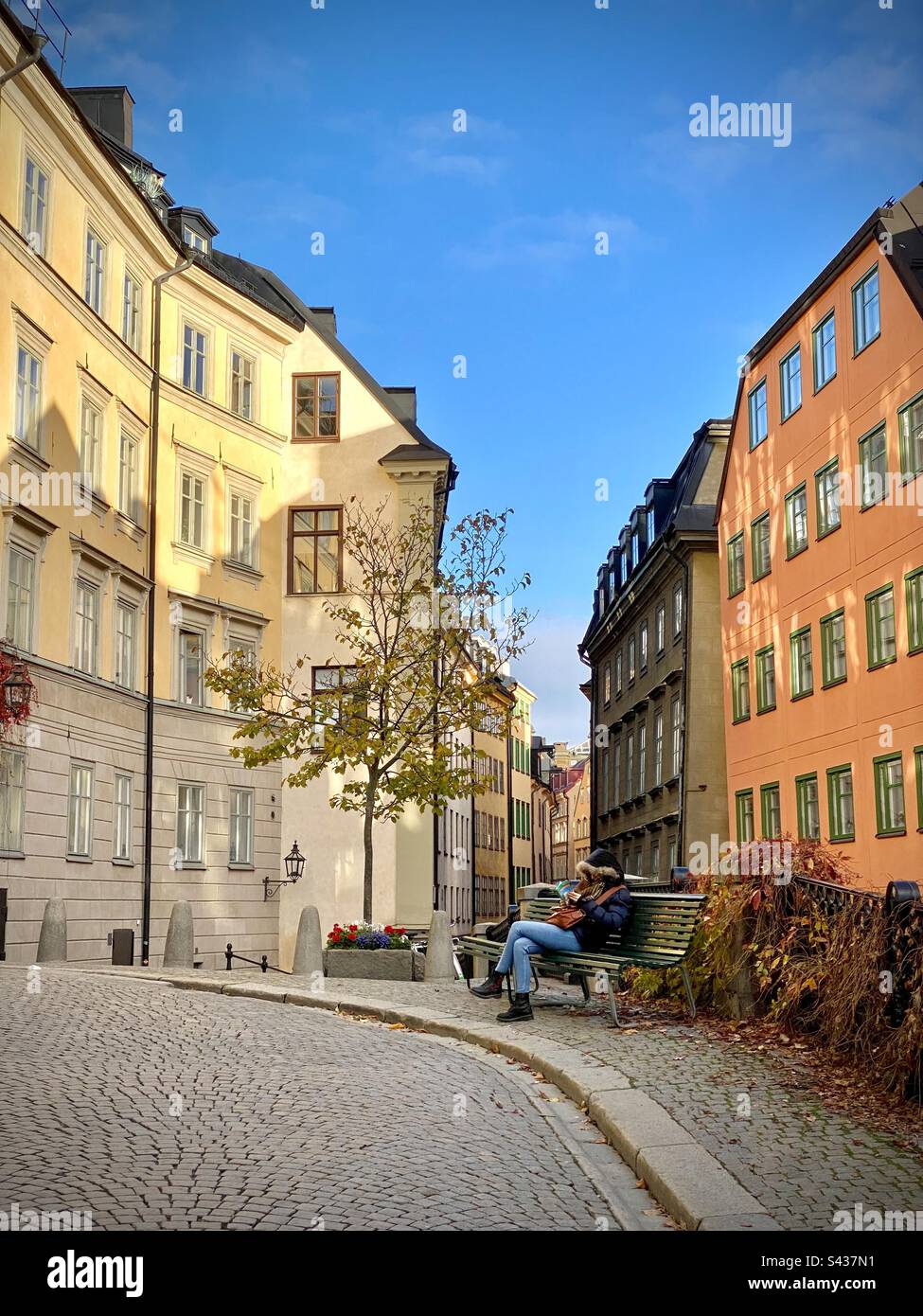 A woman sitting reading in a quiet square among the beautiful shadowy orange and yellow painted exteriors of traditional buildings on the narrow streets of Stockholm Old Town Gamla Stan in Sweden - Smartphone Captured Stock Image