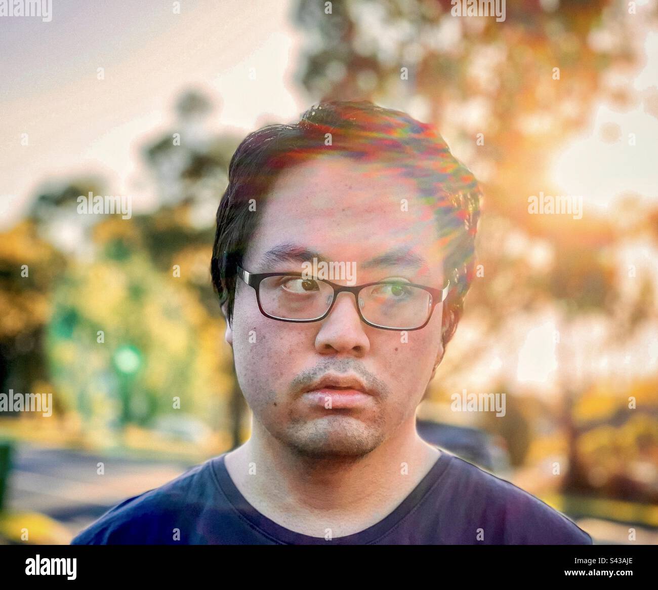 Close-up portrait of young Asian man in eyeglasses against trees and setting sun with rainbow colored lens flare effect and backlighting. - Smartphone Captured Stock Image