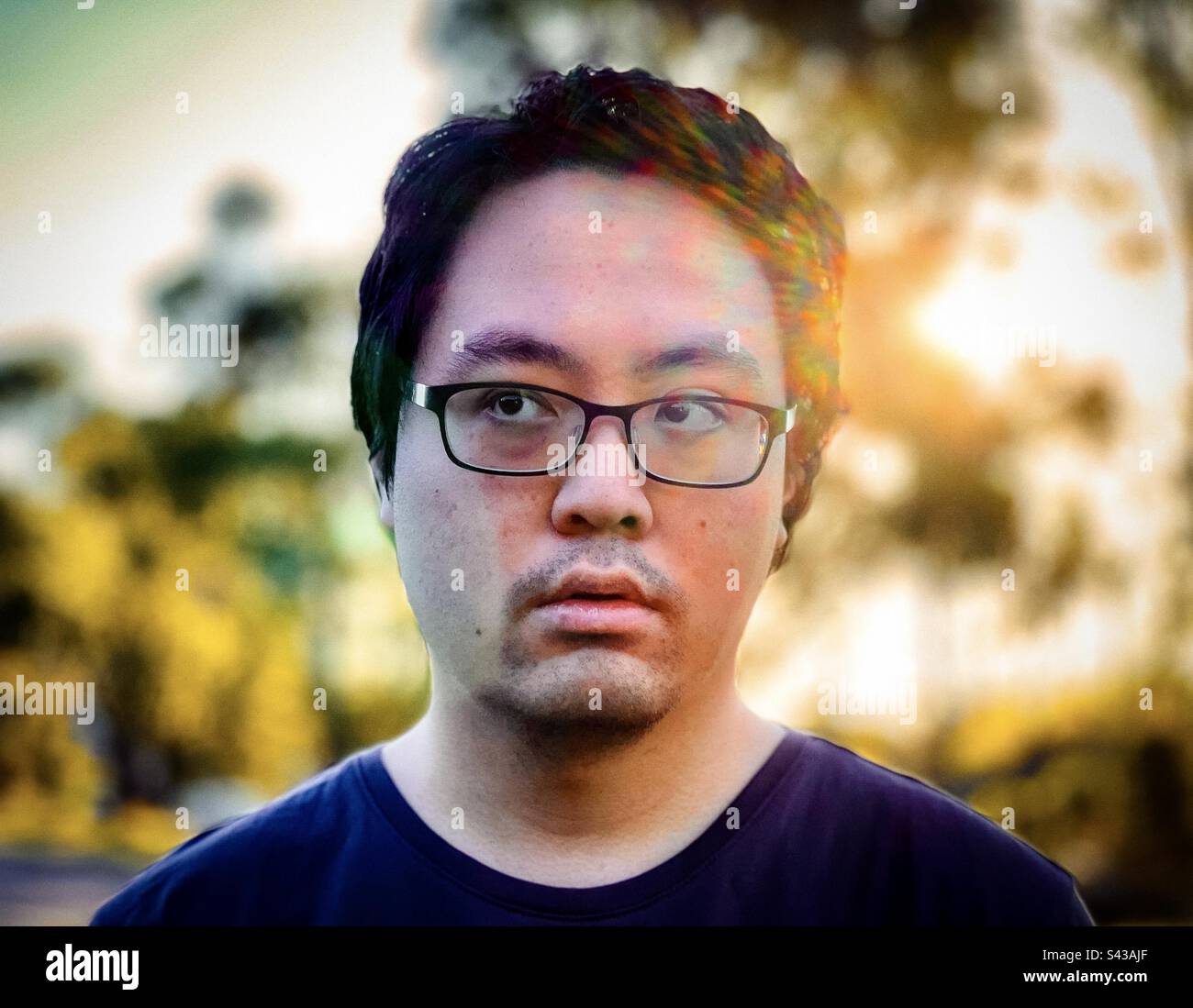 Close-up portrait of young Asian man in eyeglasses against trees and setting sun with rainbow colored lens flare effect and backlighting. - Smartphone Captured Stock Image