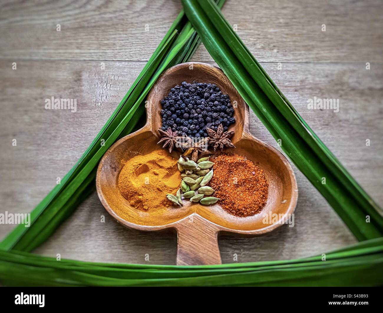 Flat lay of colorful spices arranged in decorative wooden bowl framed by green pandanus leaves also known as pandan or screw palm leaves on wooden table. Stock Photo
