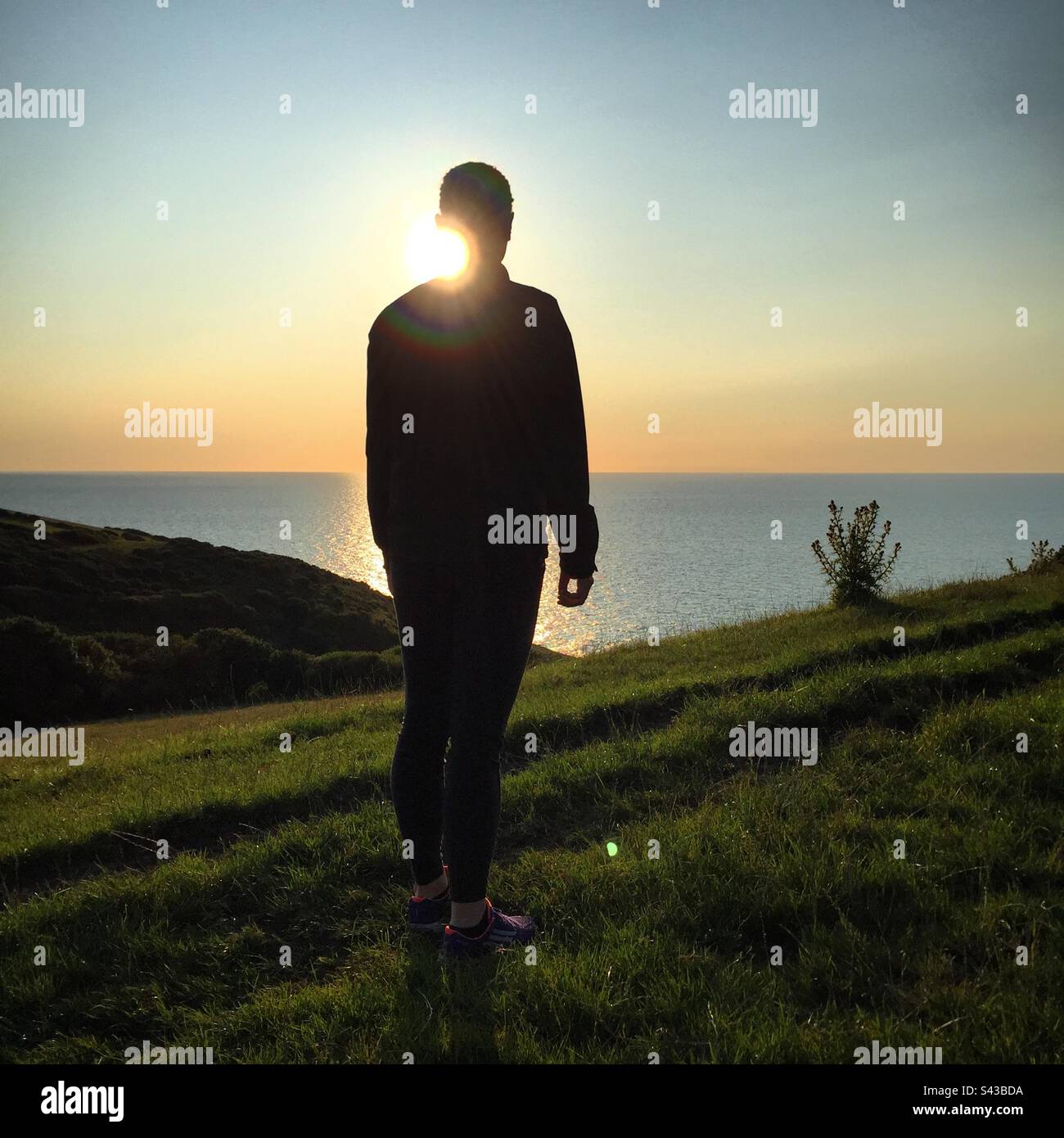 A woman in silhouette at sunset walking above the Irish Sea on a dramatic section of the popular Ceredigion Coast Path near Llangrannog in West Wales - Smartphone Captured Stock Image