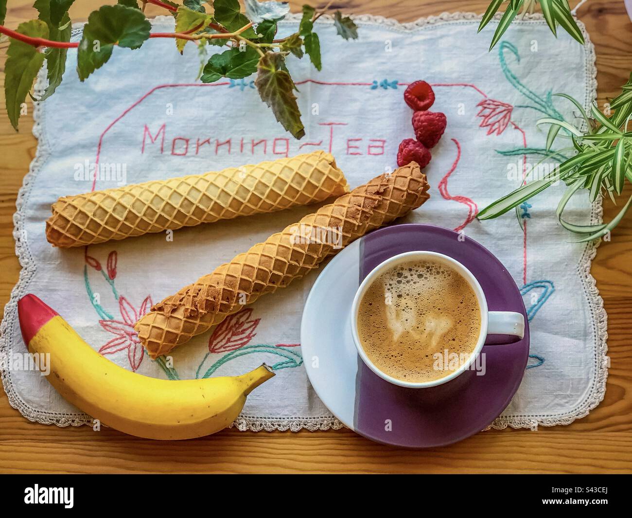 Flat lay arrangement of a banana, two crispy, wafer rolls, raspberries and a coffee cup and saucer on table cloth embroidered with the words, morning tea. - Smartphone Captured Stock Image