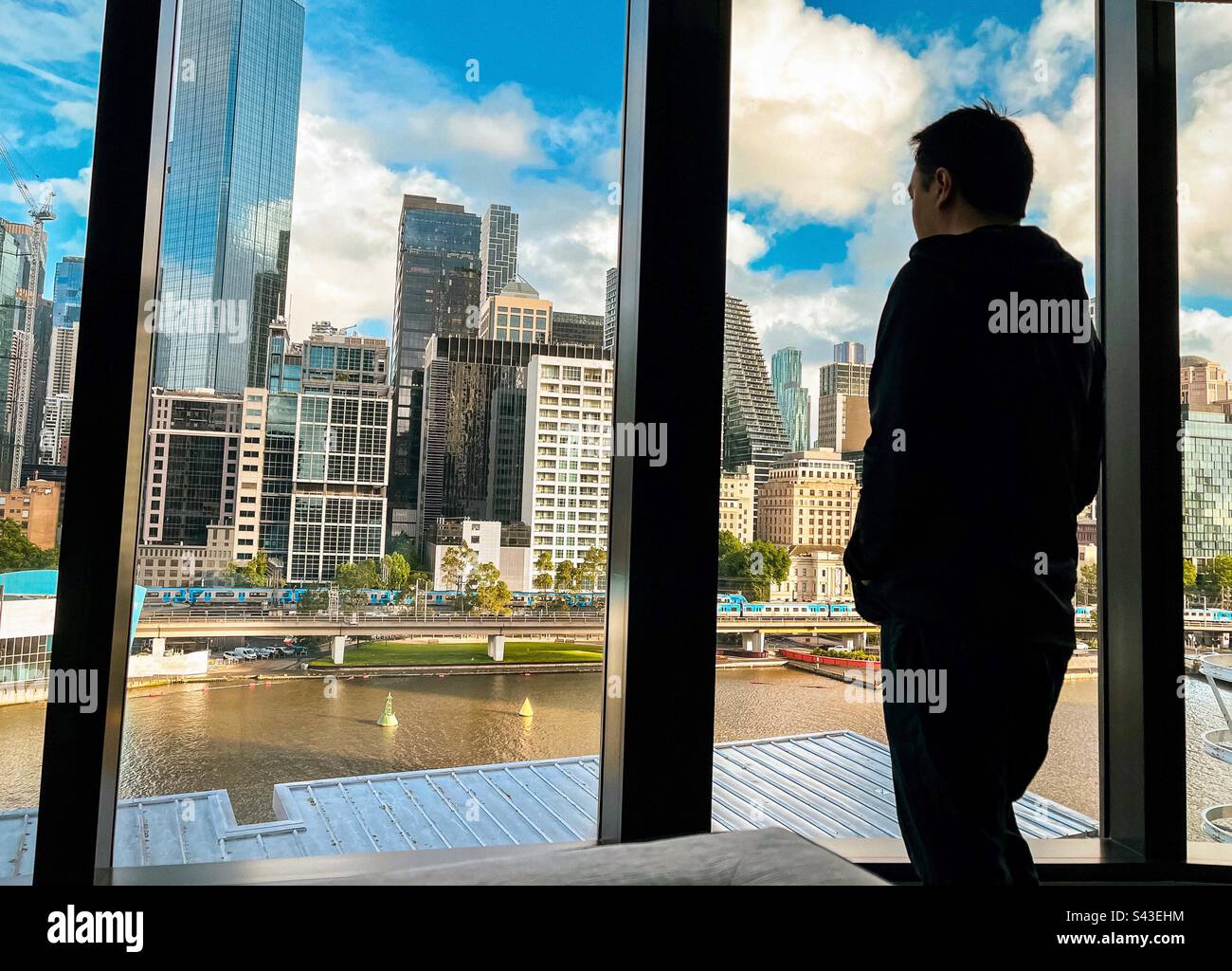 Rear view of a man standing at window view of river, city buildings, skyscrapers and urban skyline. - Smartphone Captured Stock Image Rear view of a man standing at window view of river, city buildings, skyscrapers and urban skyline. - Smartphone Captured Stock Image