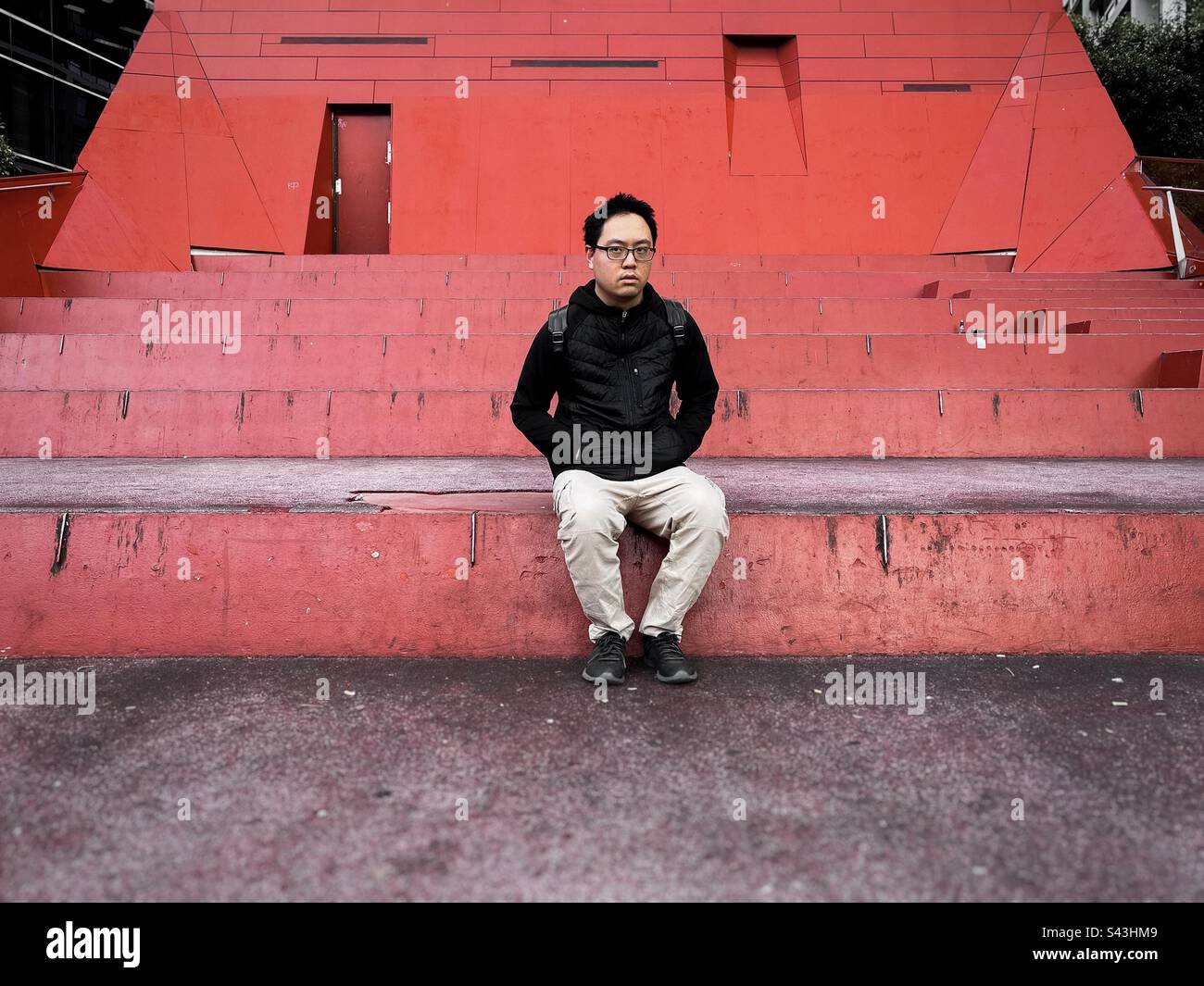 Young Asian man in eyeglasses sitting on orange steps against orange wall in Melbourne, Victoria, Australia. - Smartphone Captured Stock Image