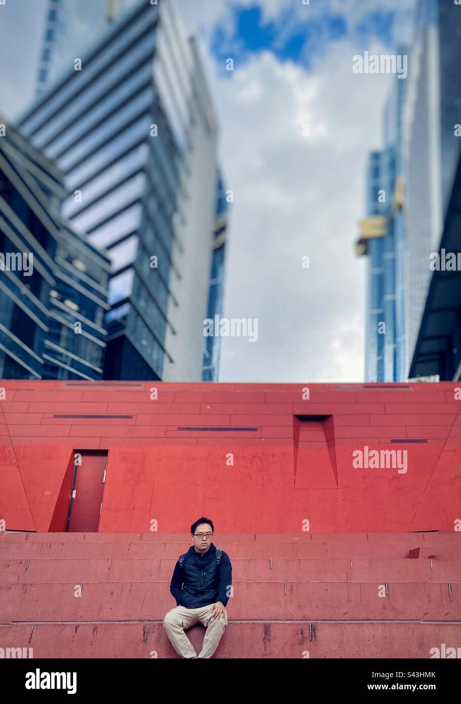 Young Asian man sitting on steps against orange wall, city skyscrapers and sky in Melbourne, Victoria, Australia. - Smartphone Captured Stock Image Young Asian man sitting on steps against orange wall, city skyscrapers and sky in Melbourne, Victoria, Australia. - Smartphone Captured Stock Image
