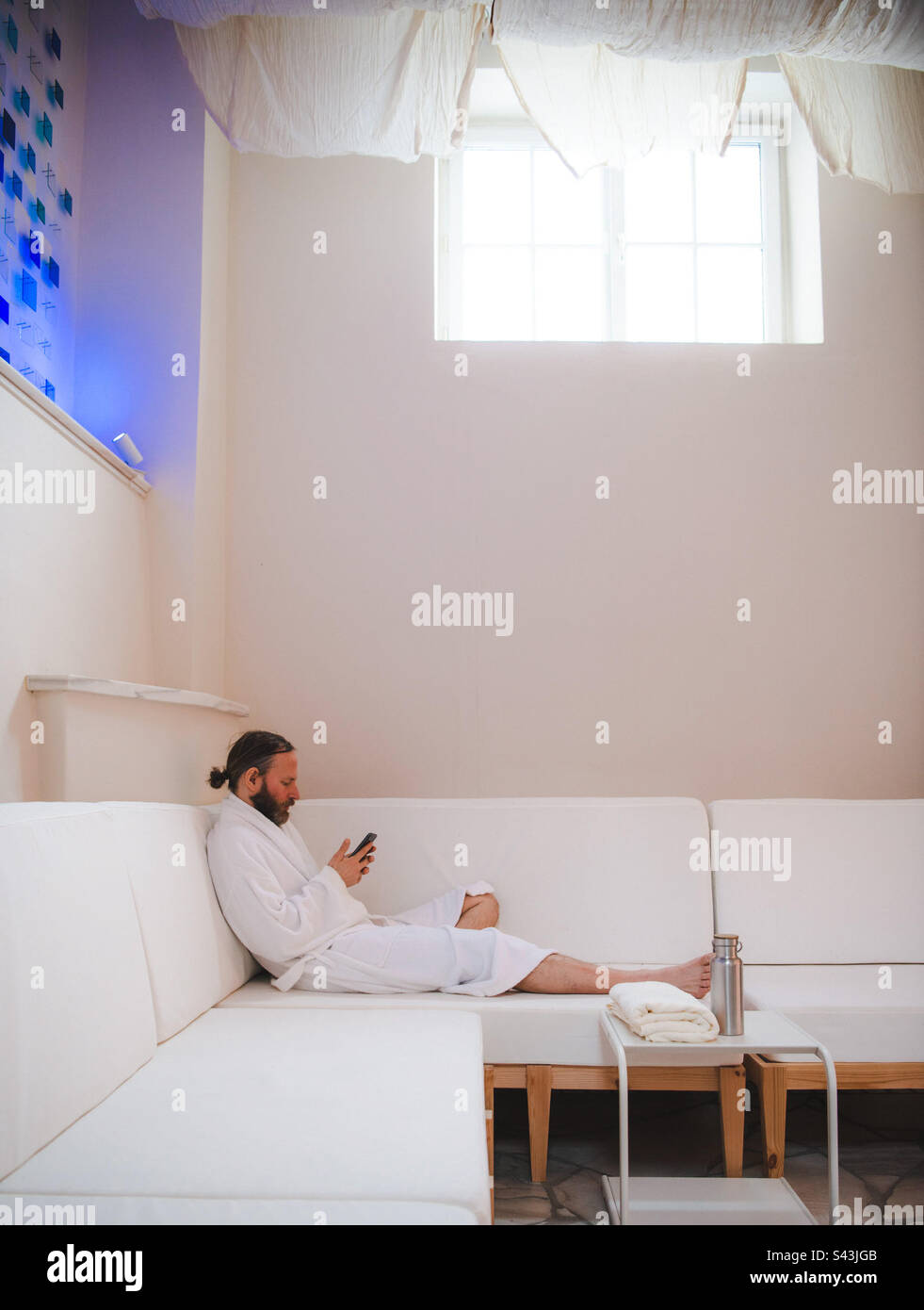 Man relaxing in robe on a white couch using his smart phone - Smartphone Captured Stock Image