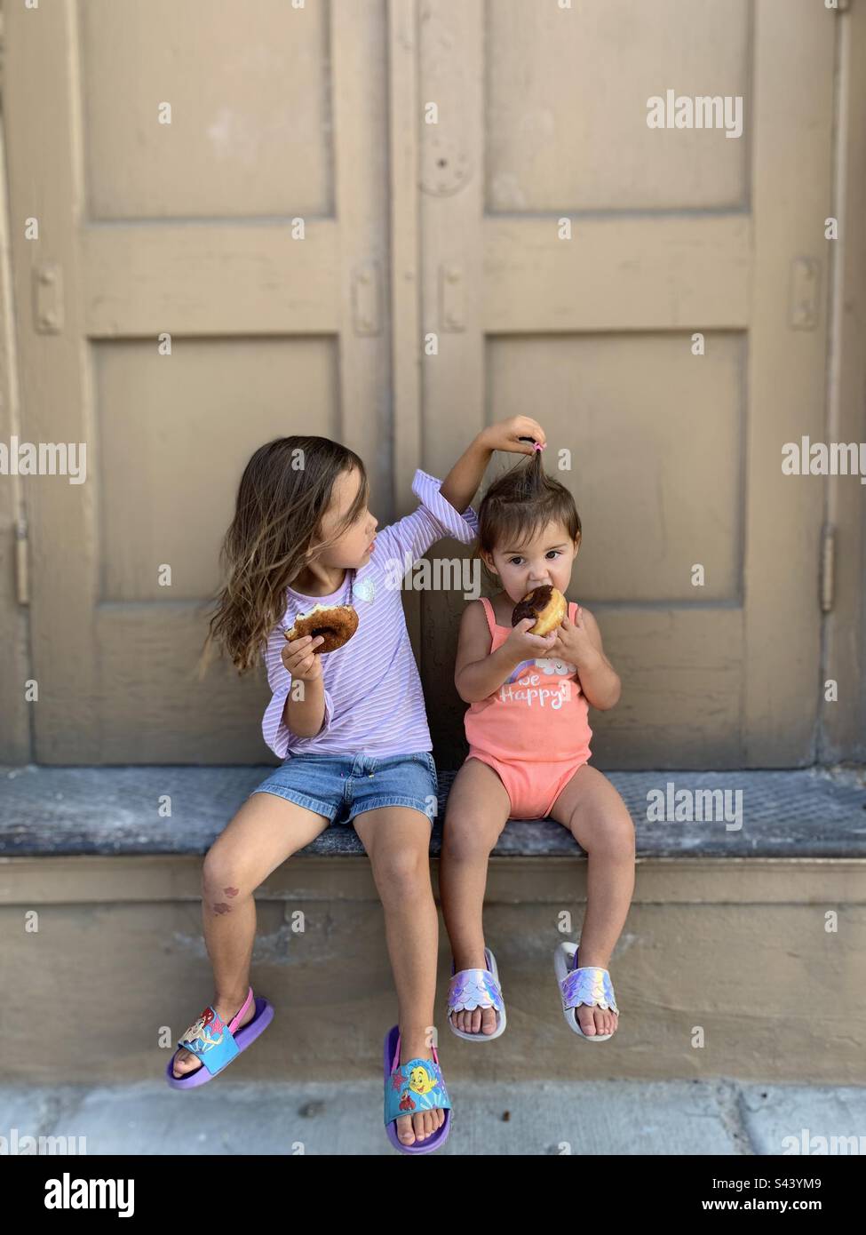 Cheeky girls eating donuts on doorstep. One is playing with the other’s hair. Stock Photo