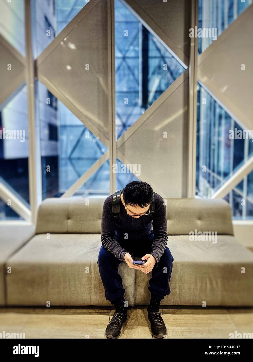 Young Asian man sitting on sofa and using mobile phone against window wall with view of city buildings. - Smartphone Captured Stock Image