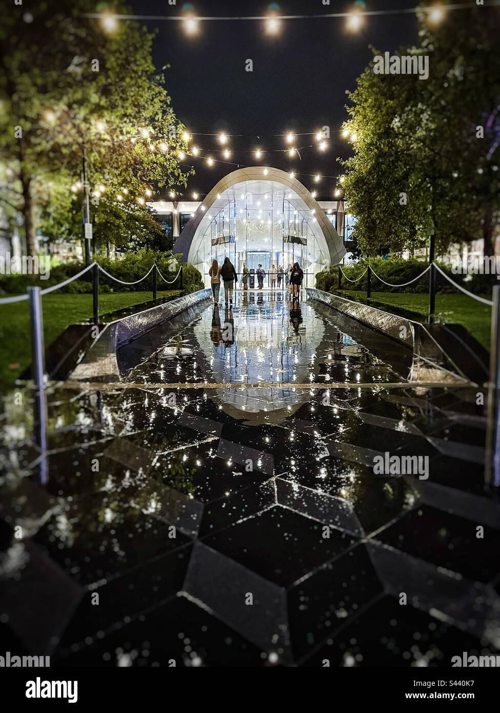 Rainy night scene at the passageway and entrance to a shopping mall illuminated with strings of lights overhead. Shoppers walking in and out. Reflections in paving tiles. - Smartphone Captured Stock Image