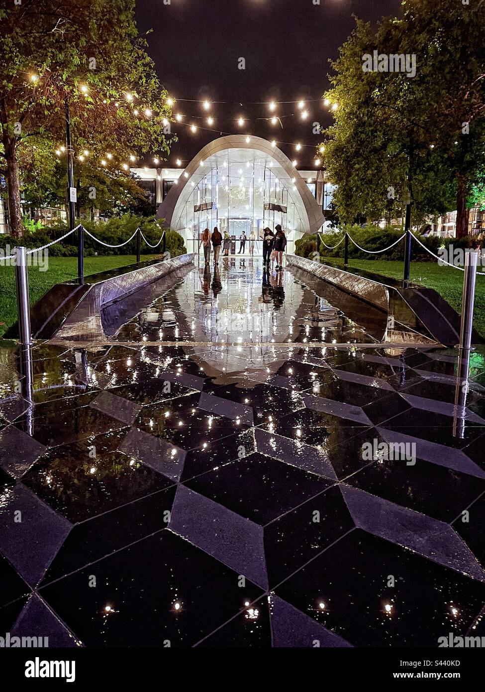 Rainy night scene at the passageway and entrance of a shopping mall. Shoppers walking in and out. Reflection in the paving tiles of passageway.String of lights hanging overhead. - Smartphone Captured Stock Image