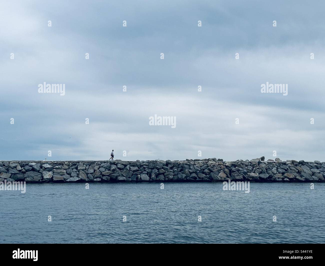 Lone figure walking along a breakwater on a cloudy da Stock Photo - Alamy