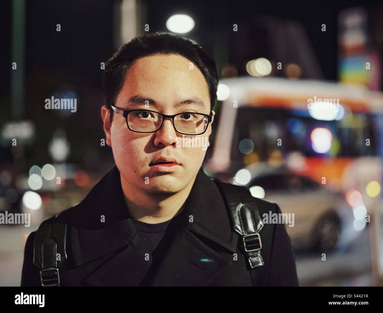 Portrait of young Asian man in eyeglasses against street traffic and street lights in the city. Retro style. Focus on foreground. - Smartphone Captured Stock Image