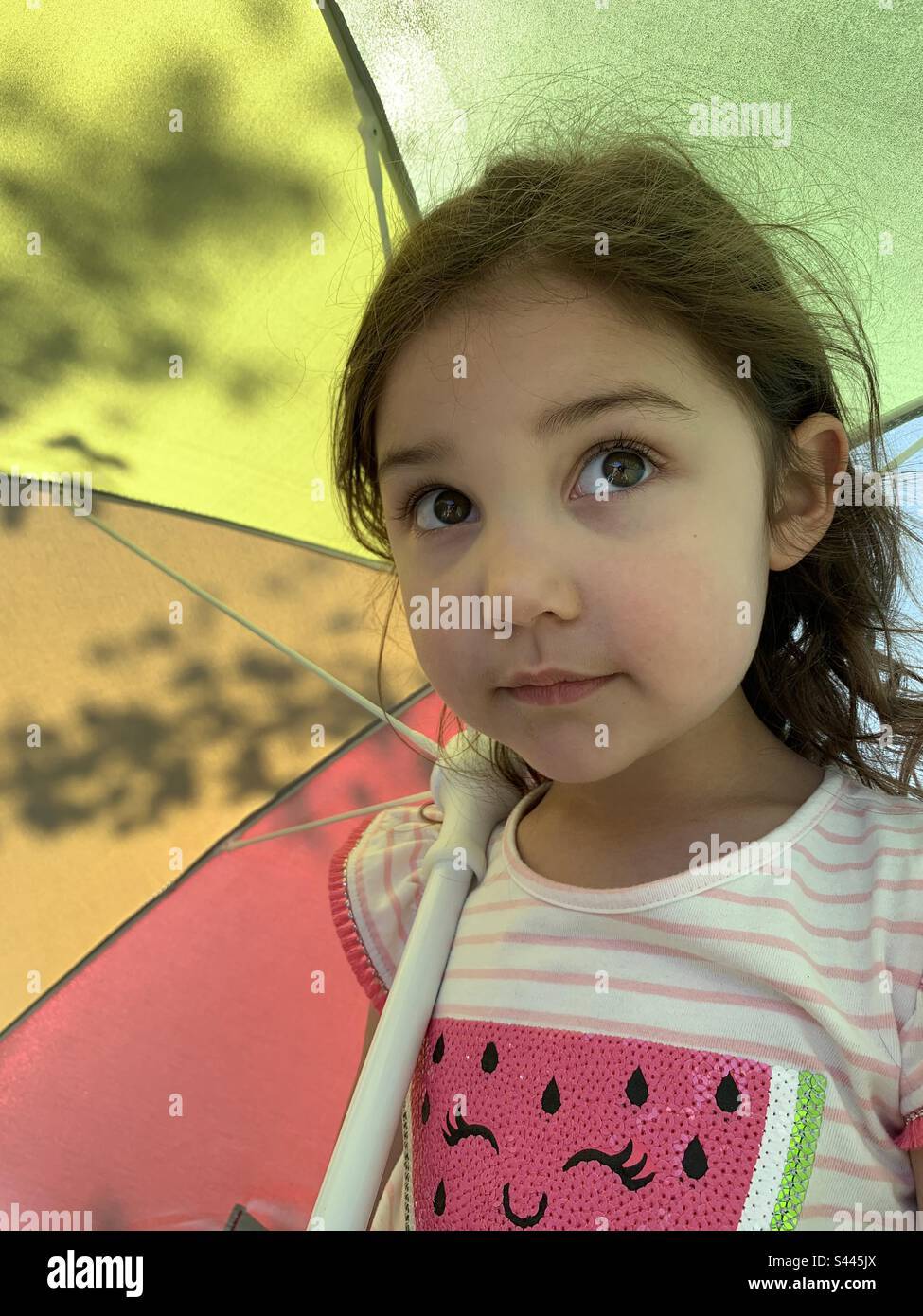 Toddler girl under rainbow umbrella in summer sun with watermelon shirt. Stock Photo