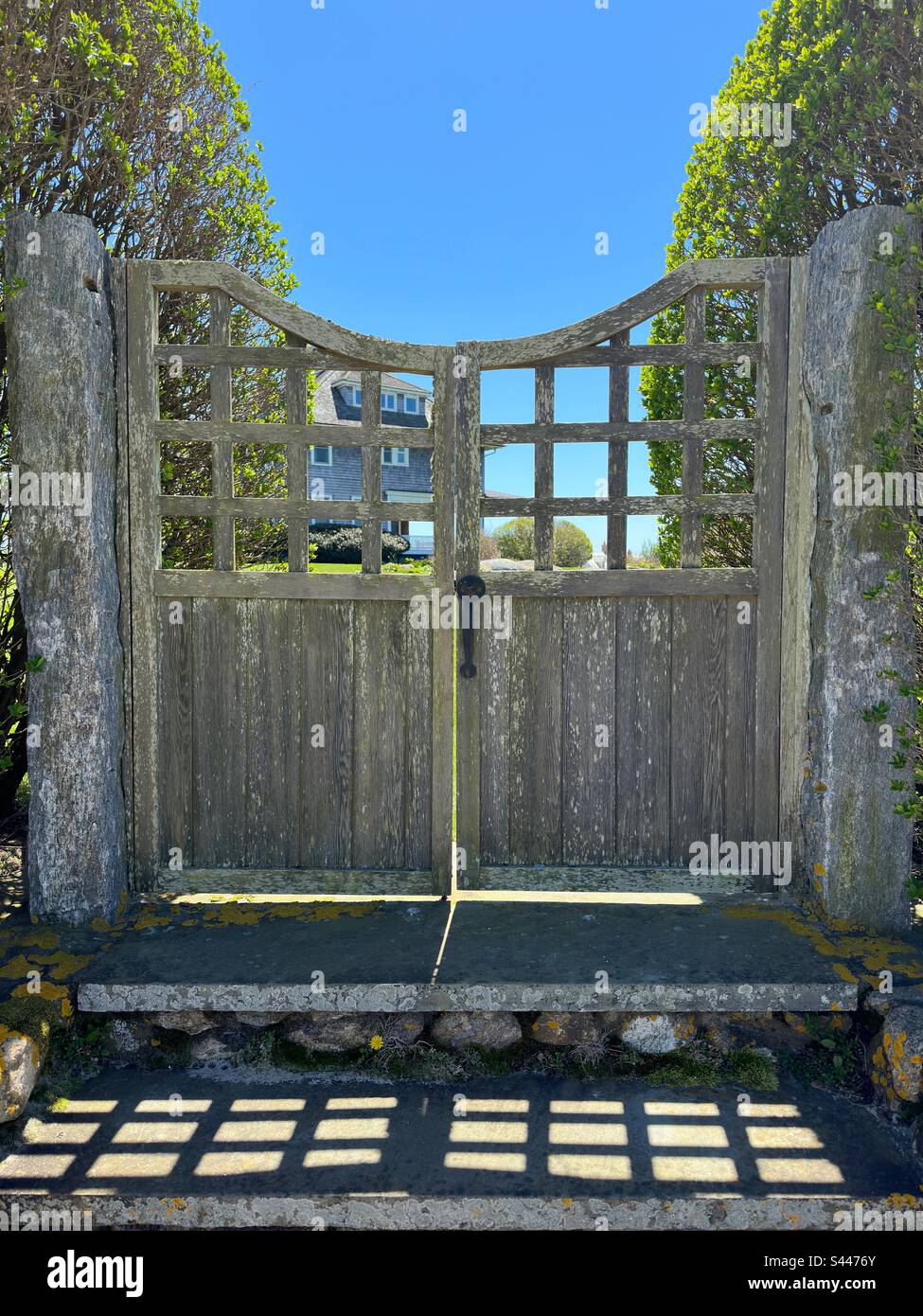 Watch Hill, Rhode Island, USA: Wooden gate with steps. Stone pillars and trees on each side that has light streaming through cracks in the gate.  House in the distance. Stock Photo