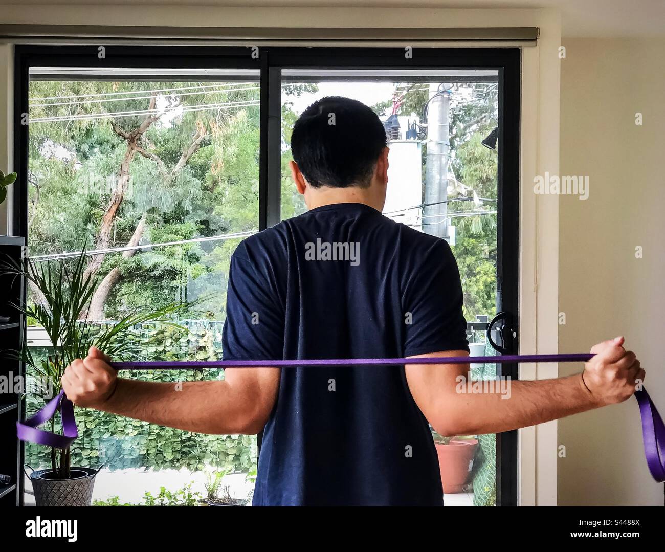 Rear view of young man using a resistance band to do strength training at home. Pulling band. Convenient at home exercise. Minimal cost. - Smartphone Captured Stock Image
