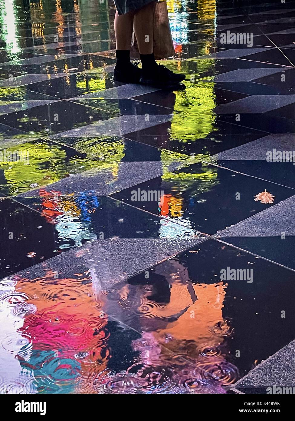Low section of man’s legs and feet standing on wet, reflective paving tiles surrounded by colorful vibrant neon light reflections on a rainy night. Art. Acidic color palette. - Smartphone Captured Stock Image