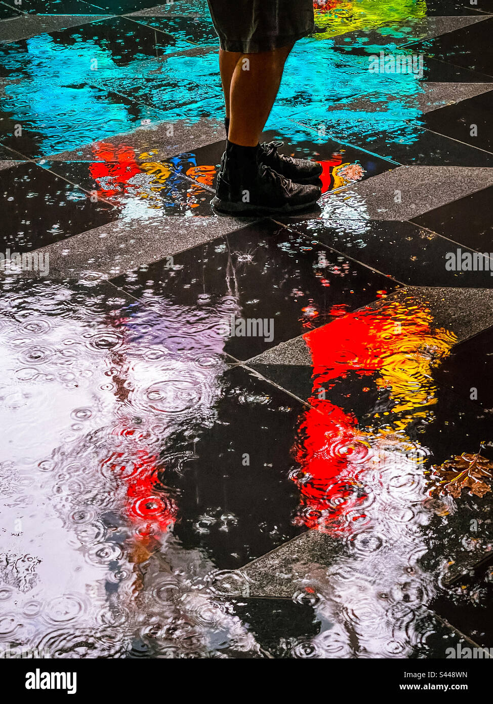 Low section of man’s legs and feet standing on wet, reflective paving tiles surrounded by colorful vivid neon light reflections on a rainy night. Art. Acidic color palette. - Smartphone Captured Stock Image