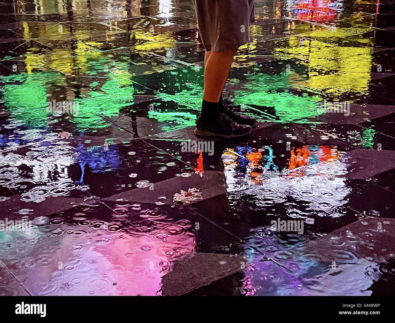 Low section of man’s legs and feet standing on wet, reflective paving tiles surrounded by colorful vivid neon light reflections on a rainy night. Art. Acidic colors palette. - Smartphone Captured Stock Image