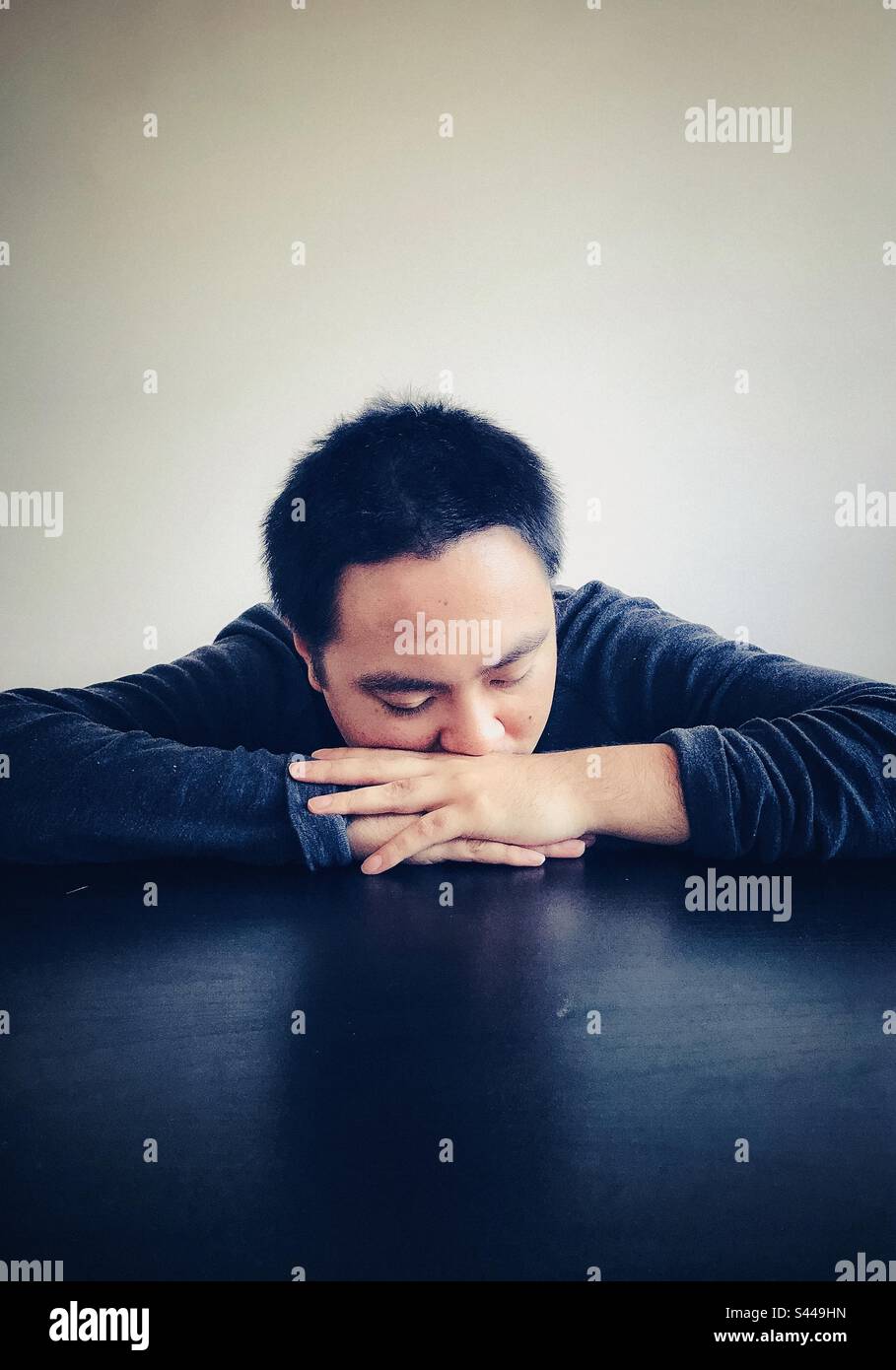Portrait of young Asian man with eyes closed and head resting on folded arms on black table. Copy space. Relaxation. Tiredness. Emotion. - Smartphone Captured Stock Image